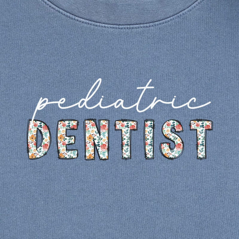 Pediatric Dentist Floral Sweatshirt in Blue Jean, close-up of the white script "pediatric" over floral "DENTIST" letters print on a folded crewneck