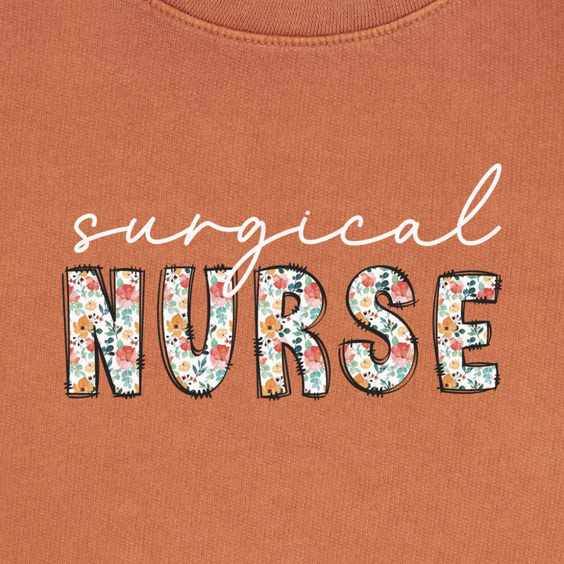 Floral Surgical Nurse Crewneck in Yam, close-up of the white script "surgical" over floral "NURSE" letters print on a folded crewneck