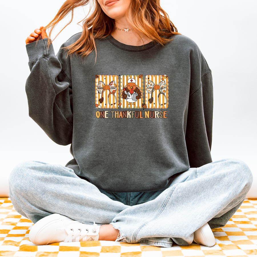 Model wearing One Thankful Nurse Thanksgiving sweatshirt in Pepper, front design of One Thankful Nurse lettering with turkey nurse, fall bows and stethoscopes, Comfort Colors 1466