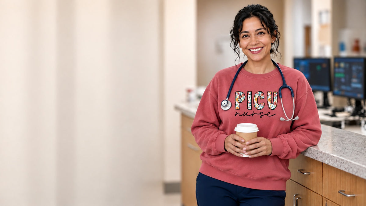 Smiling nurse leaning on a nurses' station counter with a coffee, wearing the PICU Nurse floral-letter sweatshirt in Crimson over navy scrubs, with a stethoscope and badge reel.
