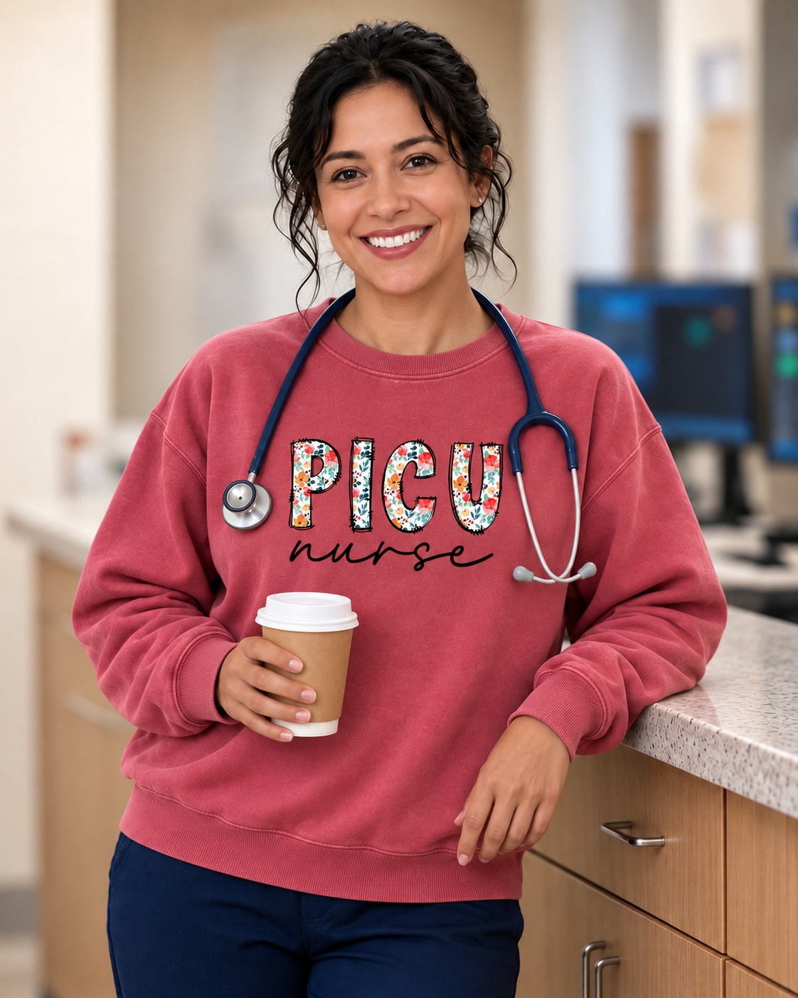 Smiling nurse leaning on a nurses' station counter with a coffee, wearing the PICU Nurse floral-letter sweatshirt in Crimson over navy scrubs, with a stethoscope and badge reel.