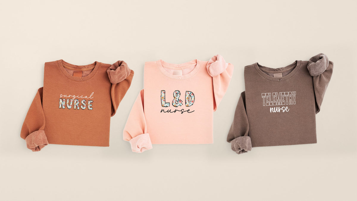 Three folded crewneck unit designs, Surgical, L&D and Telemetry, laid out on warm oat