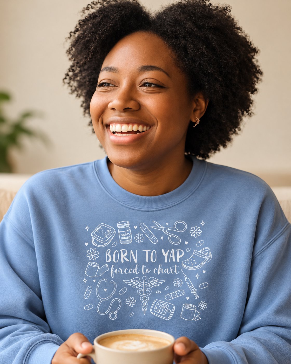 Smiling woman with a latte wearing the Born to Yap, Forced to Chart nurse sweatshirt in Blue Jean.