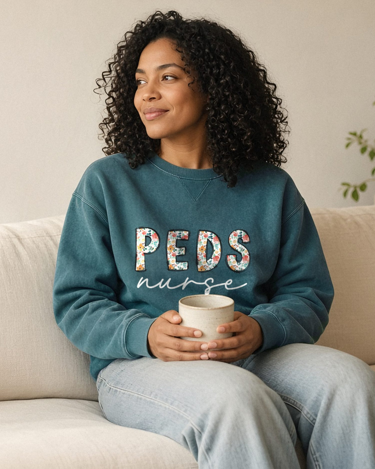 Woman on a cream sofa holding a mug, wearing a Blue Spruce PEDS nurse floral crewneck with white script