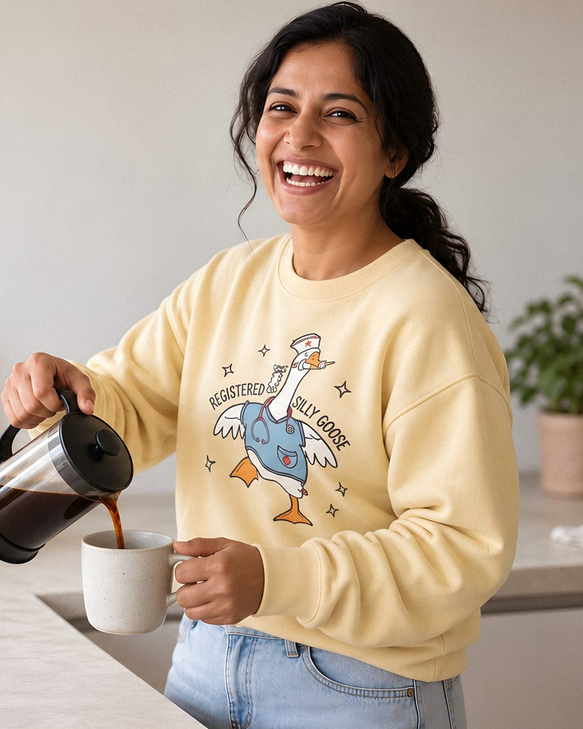 Smiling woman pouring French press coffee in a bright kitchen, wearing the Registered Silly Goose nurse sweatshirt in Butter.