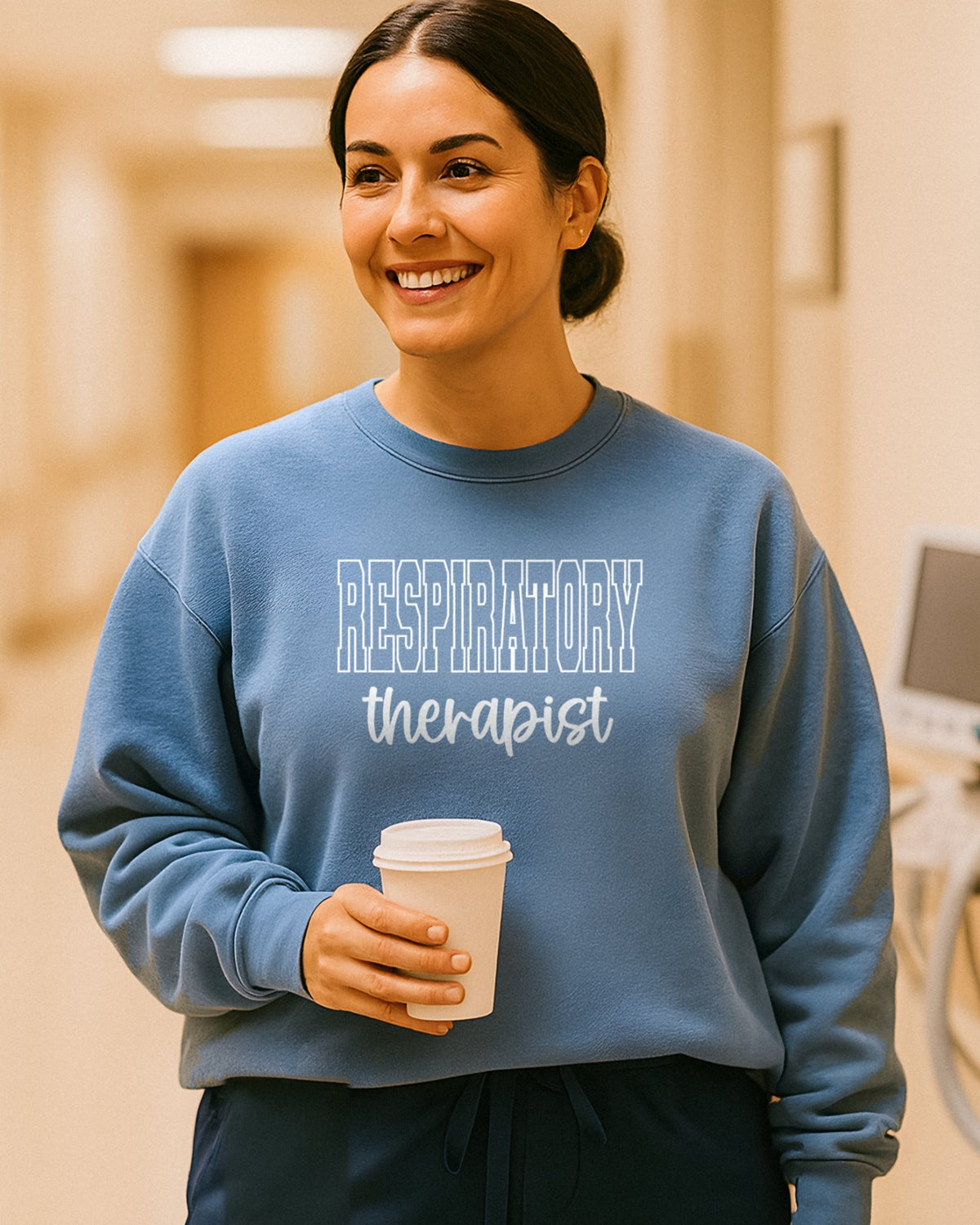 Smiling respiratory therapist in a Blue Jean Respiratory Therapist varsity crewneck over navy scrubs, holding a coffee in a warm hospital hallway beside a ventilator and wall oxygen flowmeter