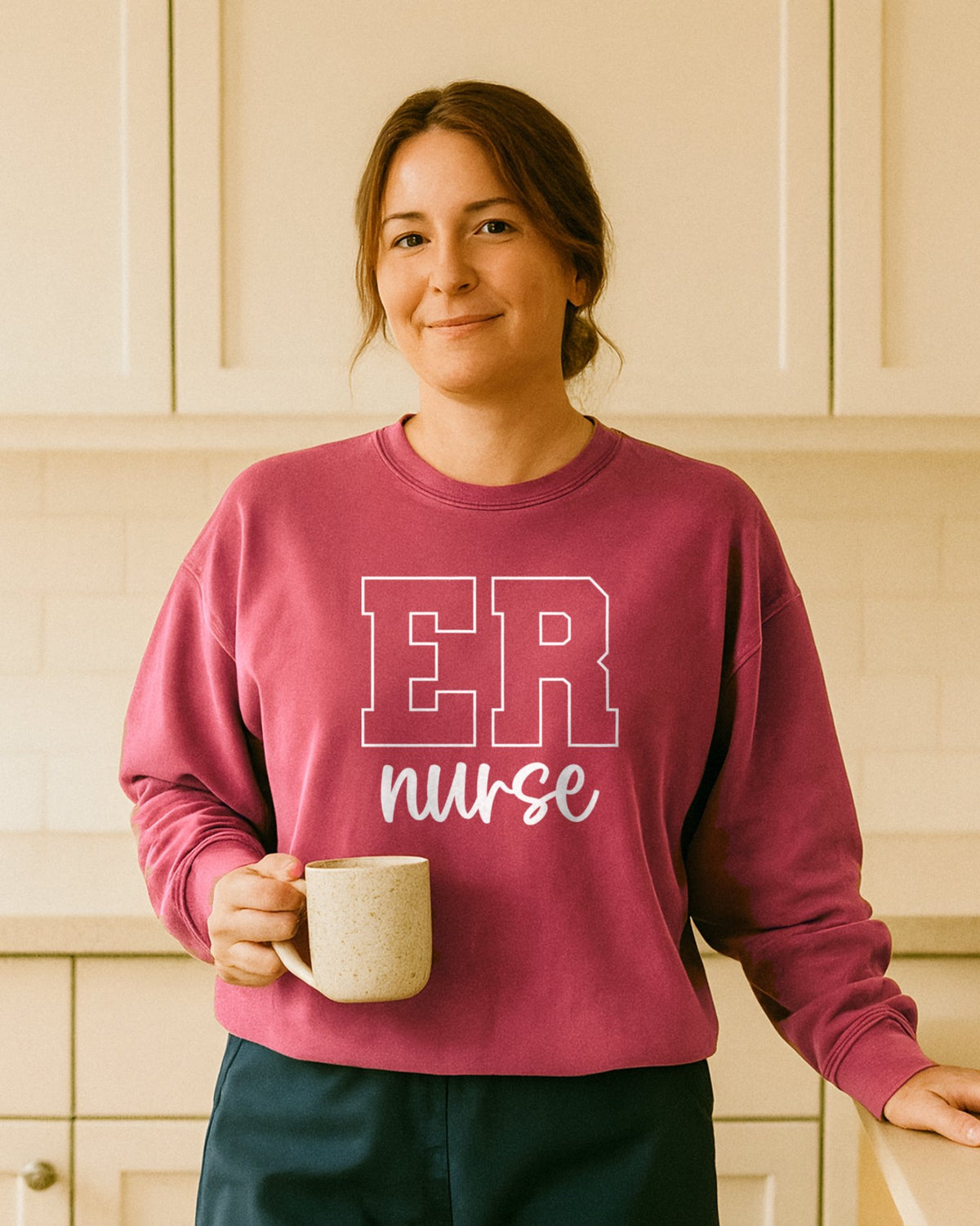Smiling nurse in a Crimson ER Nurse varsity crewneck leaning against a cream kitchen counter, holding a speckled coffee mug at her side in warm morning light