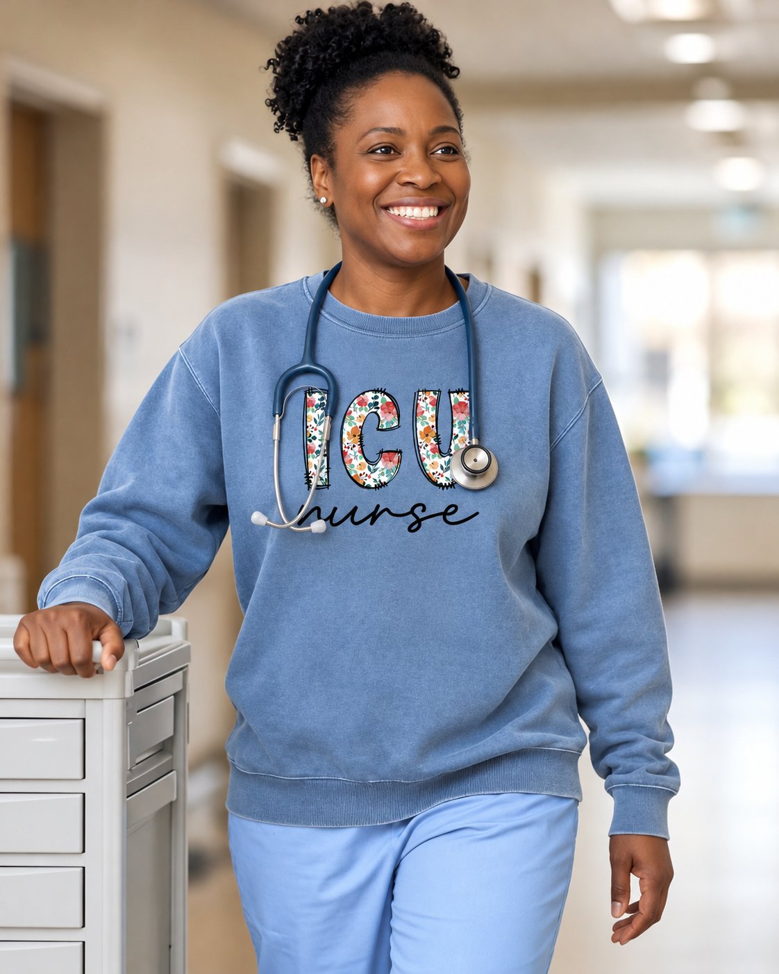 Nurse in a Blue Jean Floral ICU crewneck with a stethoscope walking beside a med cart in a bright hospital hallway